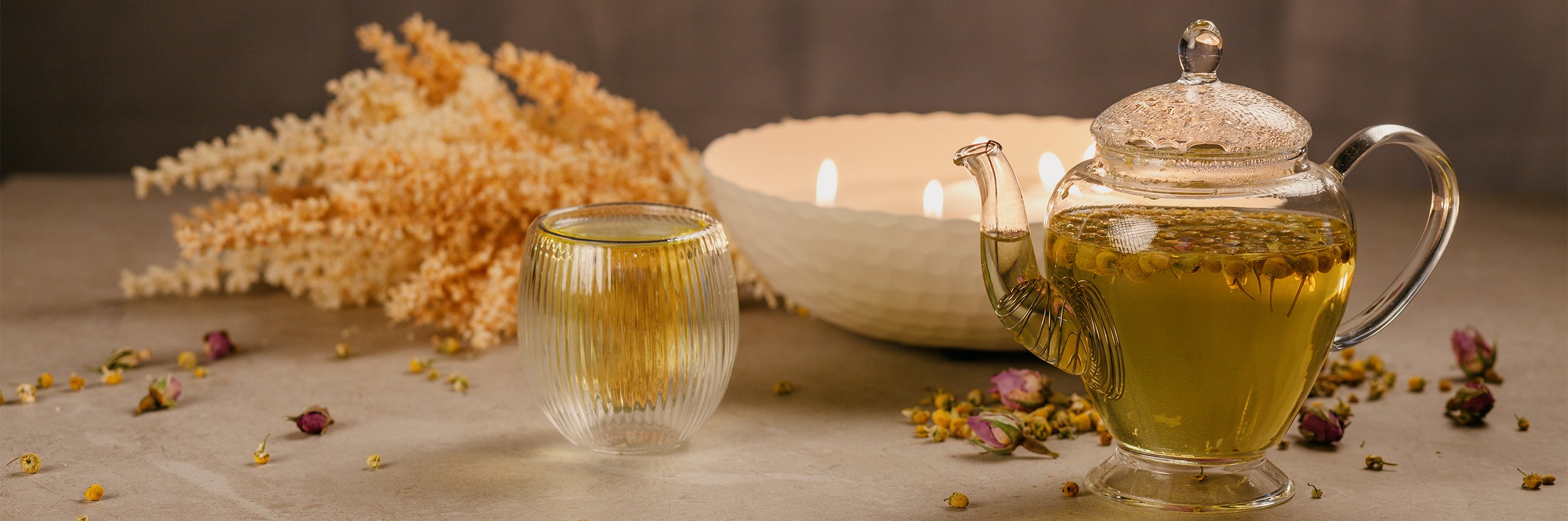 A glass teapot full of chamomile tea , next to a glass cup full of chamomile tea. There are tealight candles in a bowl in the background and some dried plants. 