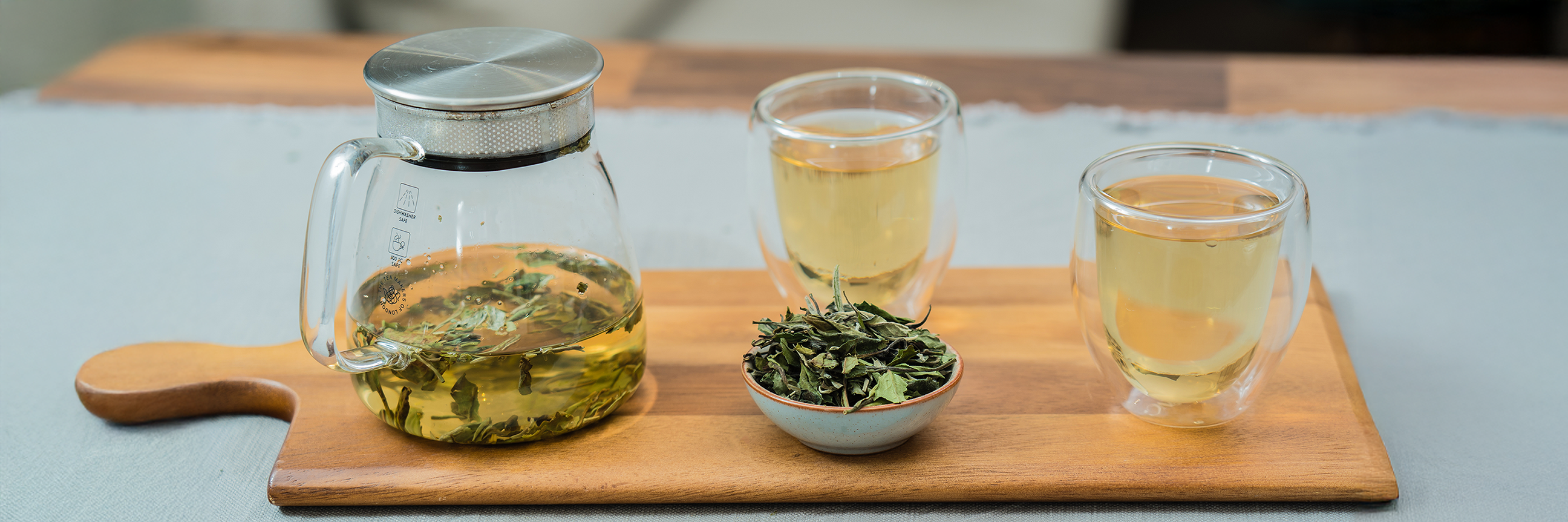 A wooden tray with a handle, with a half full glass teapot, two glasses of tea and a small bowl full of tea leaves. 