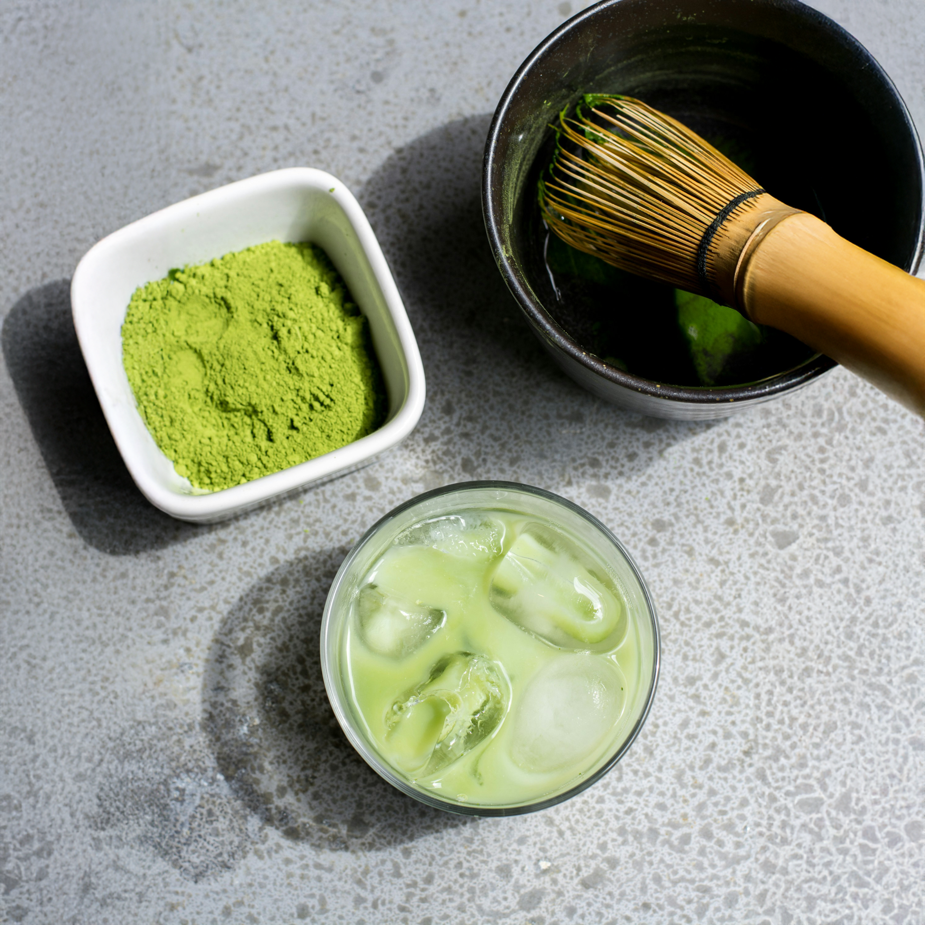 An Iced Matcha Latte, with a matcha bowl, matcha whisk and powder on a grey marble surface. 